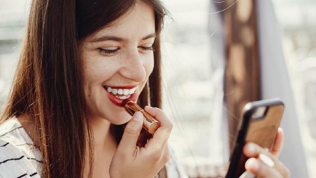 Beautiful stylish young woman applying red lipstick on lips and looking at phone screen. Happy hipster girl in retro dress relaxing and smiling on roof in sunny  european city