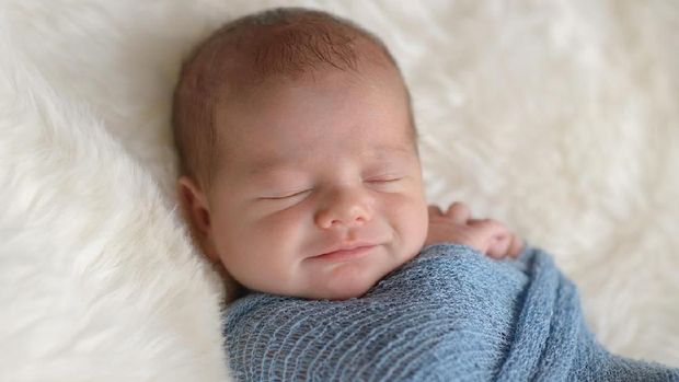 Sleeping, nine day old, newborn baby boy with a smile on his face. He is swaddled in a light blue wrap. Shot in the studio on a white, sheepskin rug.