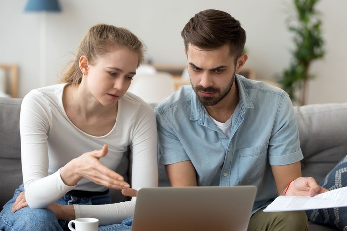 Focused man and woman sit on couch using laptop taking care of financial bills, husband and wife manage bank documents, reading regulations online, upset couple consider received papers