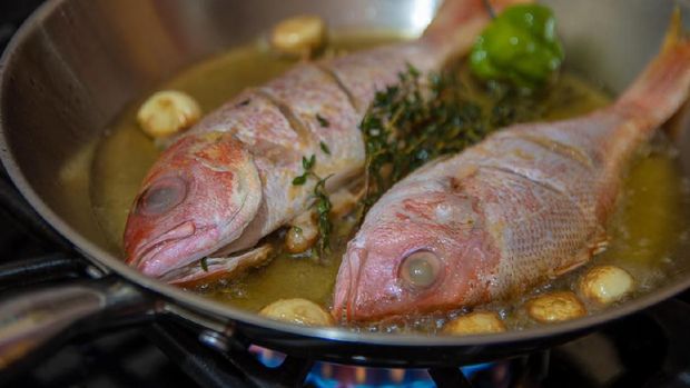 Red snapper fish being fried in frying pan on stove. Cooking vegetarian meal.