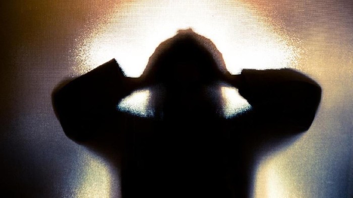 Colour backlit image of the silhouette of a woman with her hands on her head in a gesture of despair. The silhouette is distorted, and the arms elongated, giving an alien-like quality. The image is sinister and foreboding, with an element of horror. It is as if the woman is trying to escape from behind the glass. Horizontal image with copy space.