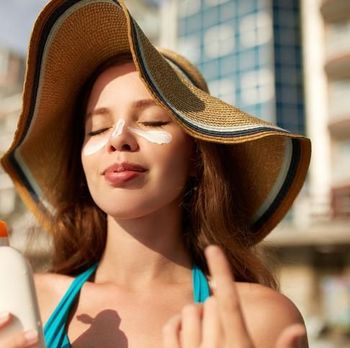Suntan lotion. Woman applying sunscreen solar cream on face. Beautiful happy cute girl puts suntan cream from plastic container bottle on her nose and cheecks. Skincare concept. Female in straw hat and bikini on beach.