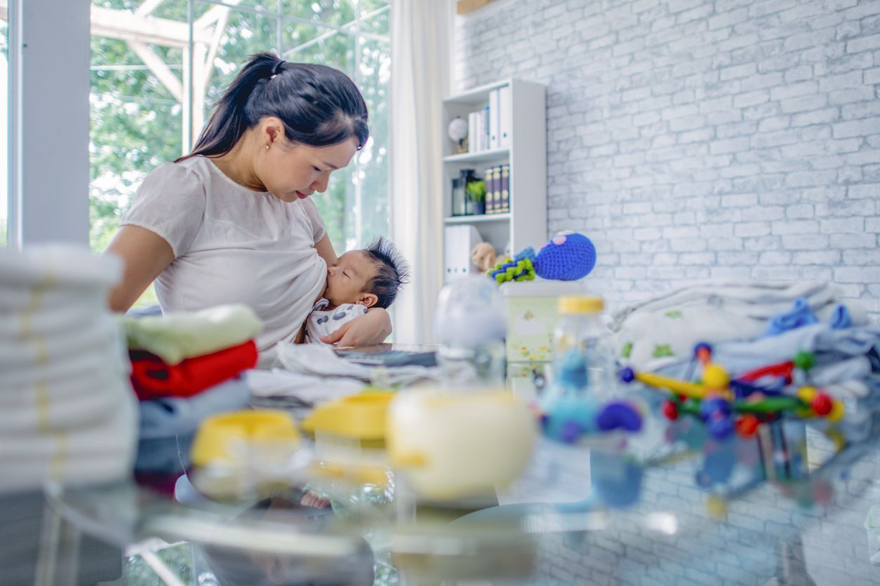 A young mom of Asian ethnicity breastfeeds her baby, while working at a table covered in baby diapers, bottles and toys.