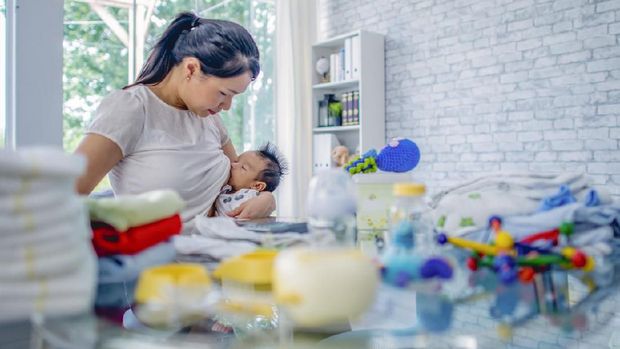 A young mom of Asian ethnicity breastfeeds her baby, while working at a table covered in baby diapers, bottles and toys.