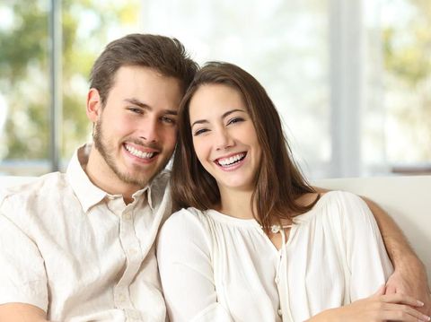 Front view portrait of a happy couple with perfect smile looking at you sititng on a couch in the living room at home in winter