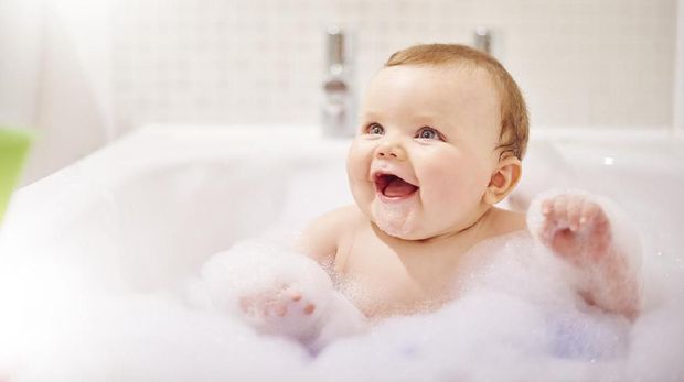 Baby boy sits in the bath waiting to have his hair washed. He is looking up and laughing to where his mother is stood next to the bath. He is sat in a baby bath seat which is hidden from view by the soap suds. The bathroom is bright and modern.