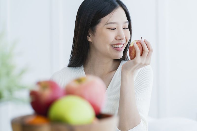 healthy beautiful attractive long black hair woman happiness smile hand hold red apple with foreground of fruit bowl white room interior background