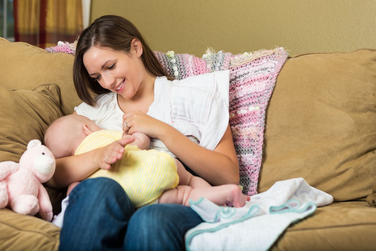 Young adult Caucasian woman is sitting on sofa at home in living room. She is breastfeeding her six month old baby daughter. Mother is smiling lovingly at baby while nursing her.