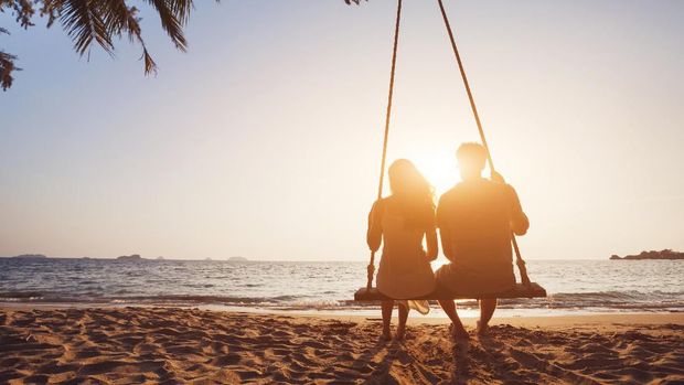romantic couple in love sitting together on rope swing at sunset beach, silhouettes of young man and woman on holidays or honeymoon