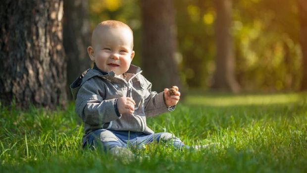 Cute funny laughing Caucasian baby boy learning to crawl, having fun playing on the lawn watching summer flowers in the garden during bright sunny day. Photo of smiling small 6 months old boy in green grass - he smiles and has fun while looking up. Happy young baby lying on tummy in nature with Beautiful little yellow duckling toys around.