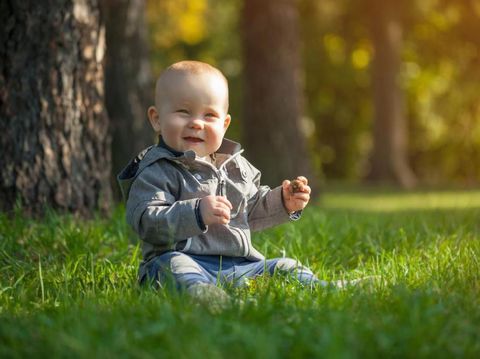 Cute funny laughing Caucasian baby boy learning to crawl, having fun playing on the lawn watching summer flowers in the garden during bright sunny day. Photo of smiling small 6 months old boy in green grass - he smiles and has fun while looking up. Happy young baby lying on tummy in nature with Beautiful little yellow duckling toys around.