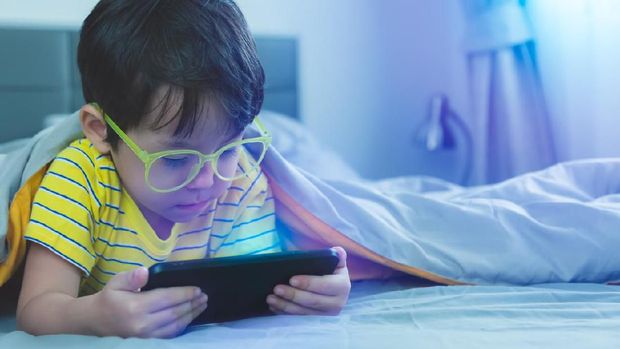Cute little child watch movie on smartphone at bed. Dangers of blue light can damage eyes. Handsome little boy can be age related macular degeneration from blue light, wear eyeglasses since childhood