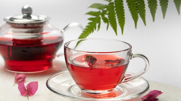 A green leaf and dried hibiscus leaves around a cup of hibiscus tea and a kettle.