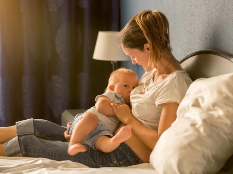 Young mother, breastfeeding on sunset on quiet evening her little baby boy in sunny living room
