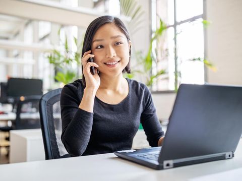 Young asian businesswoman sitting at her desk with laptop talking on mobile phone. Female professional working in modern office.