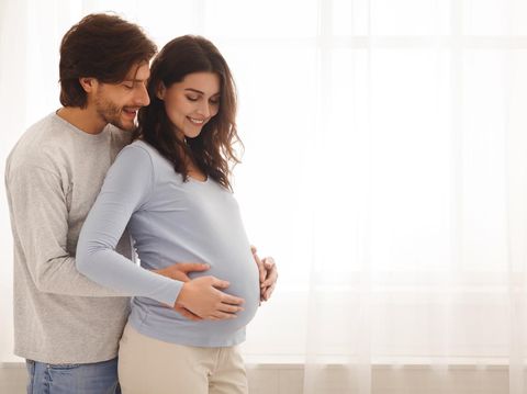 Future parents. Happy expecting couple hugging and smiling while standing near the window at home, free space