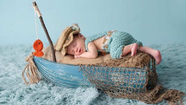 Portrait of a three week old newborn baby boy. He is sleeping in a miniature boat and wearing crocheted overalls and a fisherman's hat. Shot in the studio on an aqua colored flokati rug.