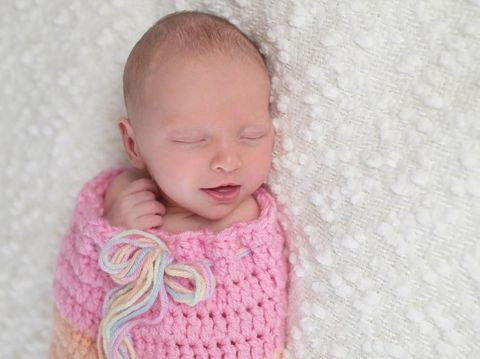 Headshot of a smiling three week old newborn baby girl bundled up in a light pink, crocheted, snuugle sack. She is lying on a white, bouncle blanket.