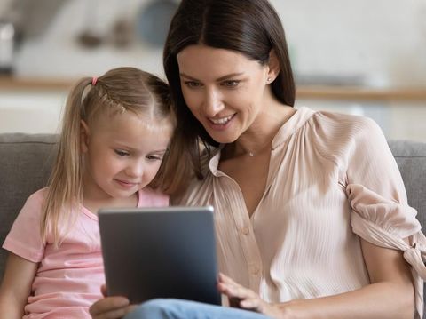 Close up mother and little daughter sit on couch having fun using tablet computer watching cartoons online or educational program. Modern wireless tech and leisure with kid, parental control concept