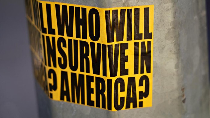 NEW YORK, NEW YORK - MARCH 22:  A view of sticker on a street lamp post reading Who will survive in America? as the coronavirus continues to spread across the United States on March 22, 2020 in New York City. The World Health Organization declared coronavirus (COVID-19) a global pandemic on March 11th.  (Photo by Cindy Ord/Getty Images)