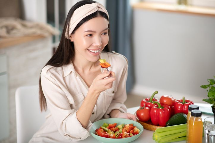 Right food. Good looking smiling young woman in a white blouse sitting at a table at home, eating with fork natural vegetable salad.