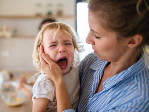 A mother holding a crying toddler daughter indoors in kitchen when cooking.