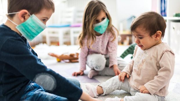 Little children wearing pollution masks at home. Two toddler kids are wearing masks while baby is crying and it’s hard to put mask on her face.
