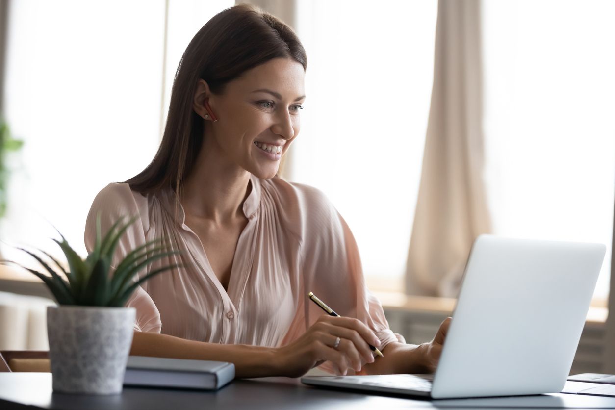 Smiling young woman wearing headphones sit at desk working at laptop making notes, happy millennial female in modern earphones study watch webinar on computer handwrite in notebook