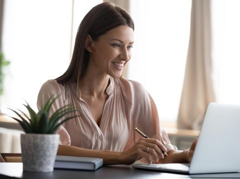 Smiling young woman wearing headphones sit at desk working at laptop making notes, happy millennial female in modern earphones study watch webinar on computer handwrite in notebook