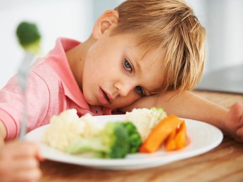A cute young boy looking at the vegetables on his table while holding a piece of broccoli on his fork