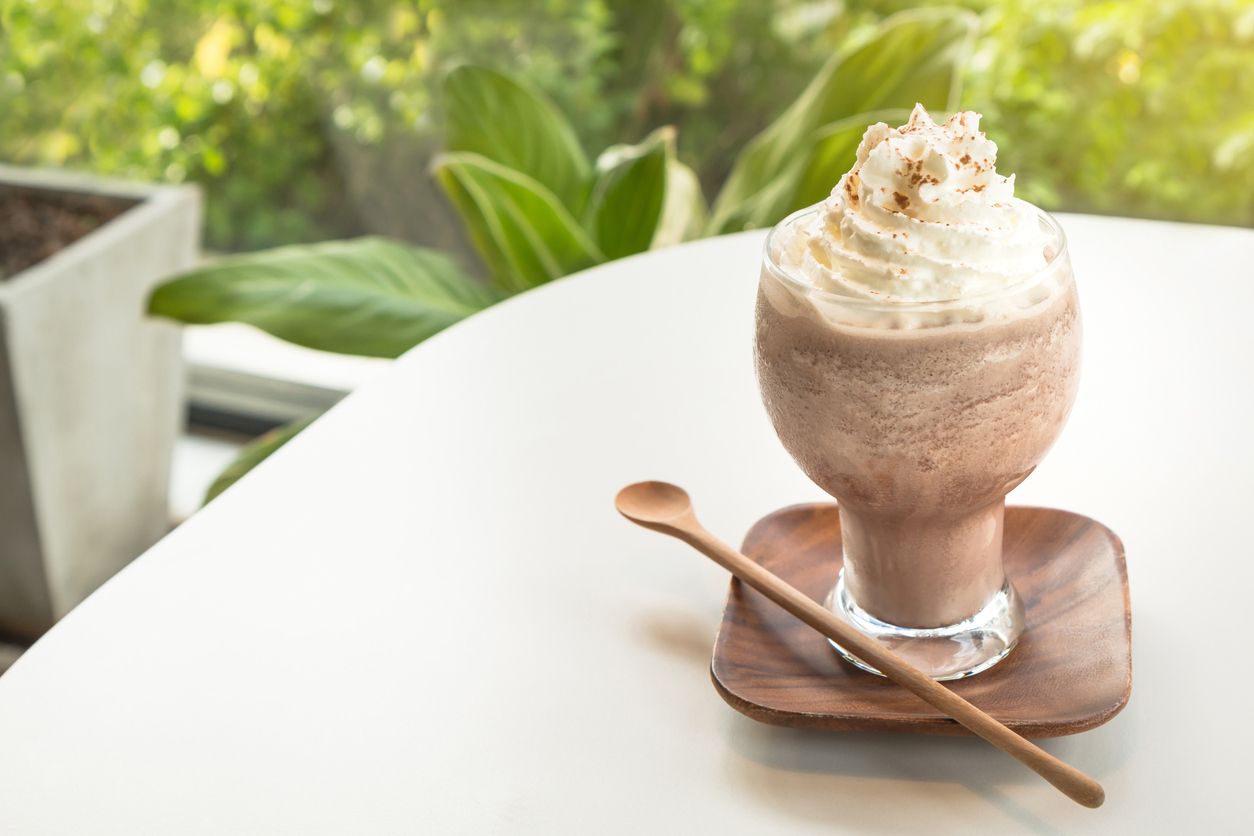 Chocolate smoothies (Cocoa blended) topped with whipped cream and cocoa powder in glass cup on white table. Wooden saucer and spoon. Interior coffee shop.