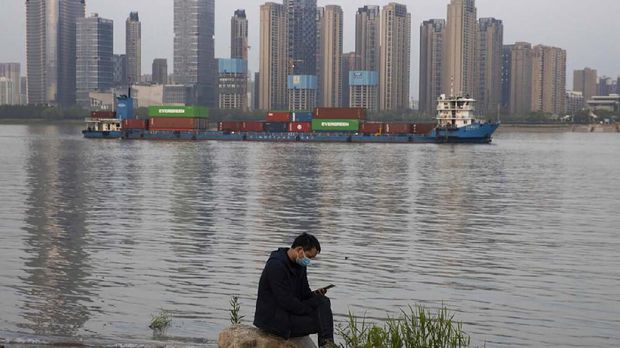 In this Monday, April 13, 2020, photo, a man wearing a mask against the new coronavirus checks his phone as a container ship cruises along the Yangtze River in Wuhan in Central China's Hubei province. China's exports fell further in March compared with February amid a global economic slowdown caused by the coronavirus pandemic, the government reported Tuesday. (AP Photo/Ng Han Guan)