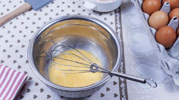Beaten eggs and a hand whisk in a stainless steel bowl on a country kitcken table with a box of eggs