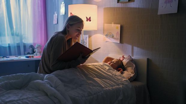 Young Loving Mother Reads Bedtime Stories to Her Little Beautiful Daughter who Goes to Sleep in Her Bed.