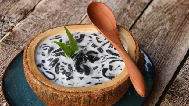 Es Dawet Ireng, the Javanese cold dessert of black cendol (tapioca short-noodle jelly) made from toasted paddy straw. It is served with crushed ice, coconut milk, and palm sugar syrup in a thick wooden bowl that has been carved out of single wooden block. A ceramic plate of solid color underlines the bowl. A wooden spoon is placed on the bowl rim. The dessert is arranged on a rustic wooden table.