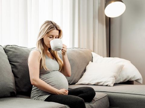 Pregnant woman sitting on her sofa drinking coffee and tea.