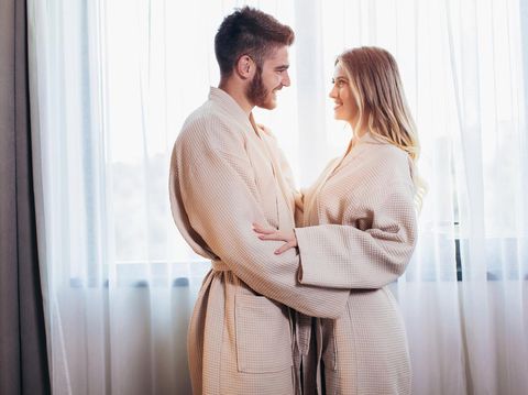 Side view of excited man is lying over woman in bed. They are looking at each other eyes and laughing