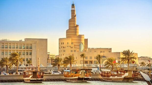 Traditional wooden dhow anchored at Dhow Harbor in Doha Bay with spiral mosque and minaret in the background at sunset. View from Corniche promenade. Qatar, Middle East, Arabian Gulf.