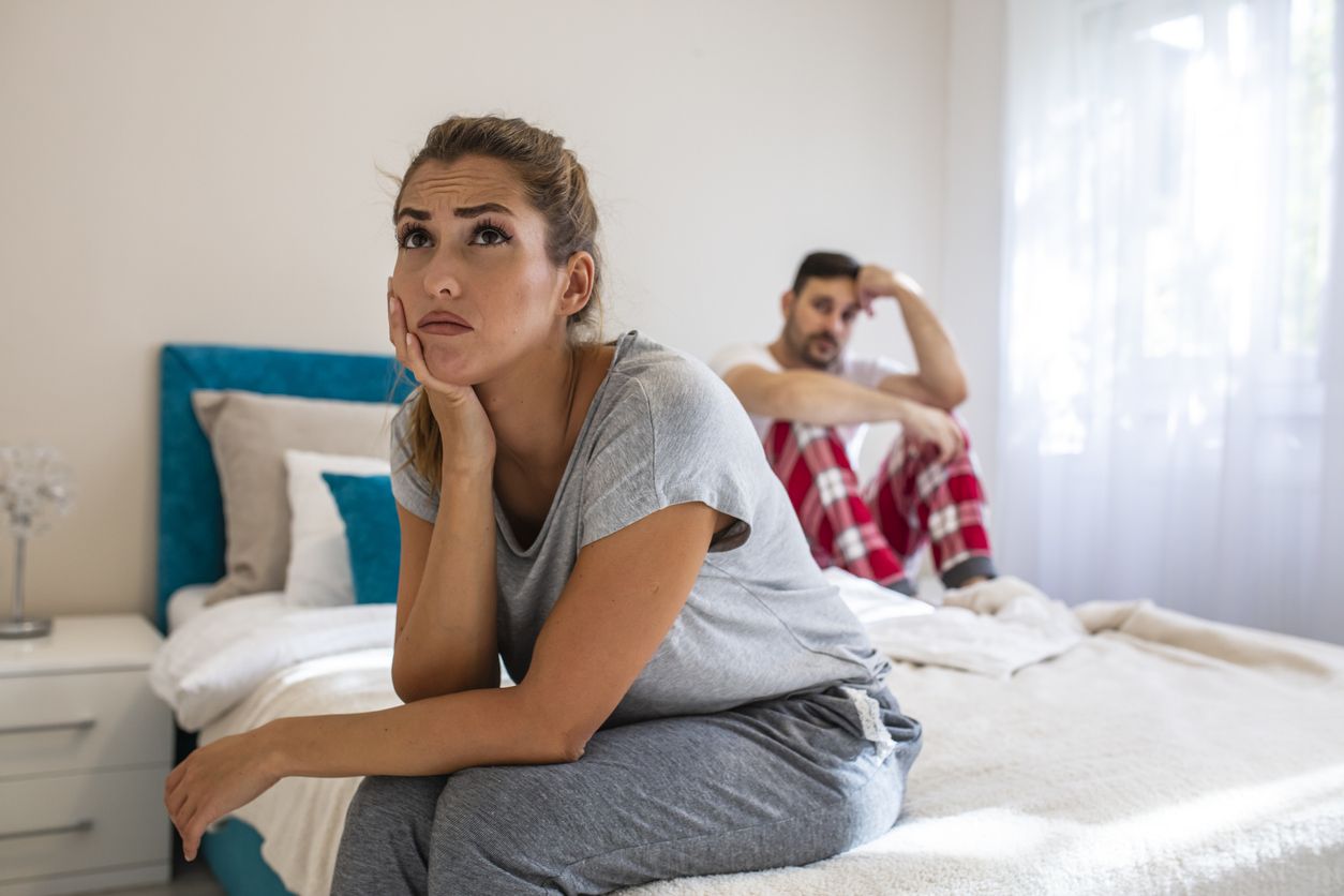 We have a problem. Young upset girl sitting on the edge of the bed, against her boyfriend, lying in bed. Frustrated sad girlfriend sit on bed think of relationship problems, thoughtful couple after quarrel lost in thoughts