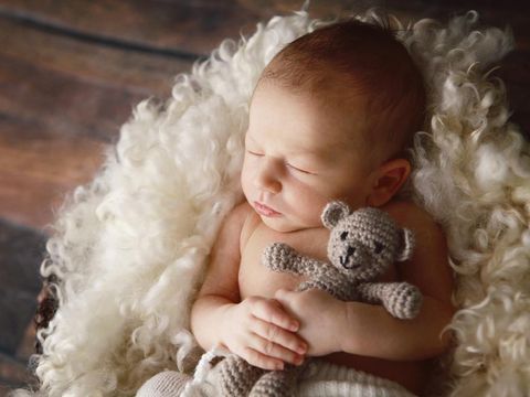 A beautiful newborn baby boy hugs a little teddy bear as he sleeps. He is lying on a soft blanket of curly wool.