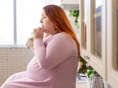 Relaxation, leisure, rest, domesticity and coziness. Beautiful charming young female with chubby cheeks and curvy body sitting on sofa barefooted, keeping one foot on floor, smiling at camera