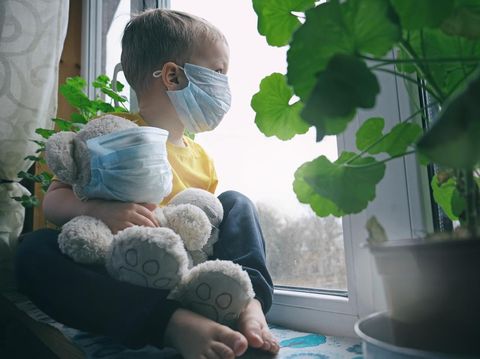 Quarantine, threat of coronavirus, virus protection, pandemic. Child and his teddy bear both in protective masks sits on windowsill inside house and looks out the window.