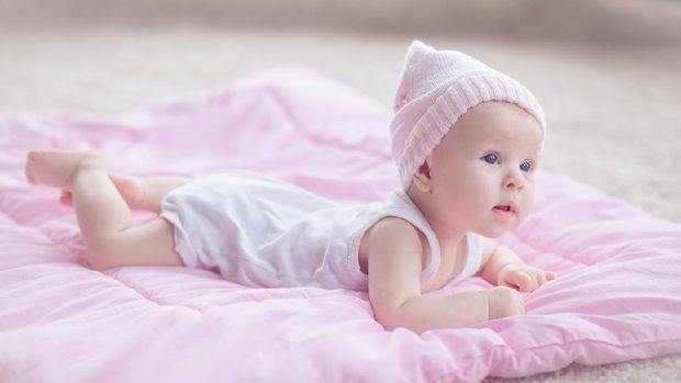 Cute playfull baby girl, wearing pink winter hat, lying at her tommy on a pink comforter on the floor. She's curious about her room interior.