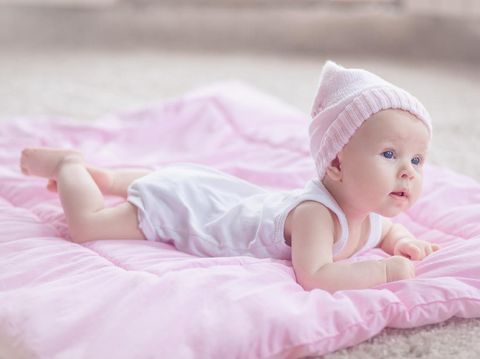 Cute playfull baby girl, wearing pink winter hat, lying at her tommy on a pink comforter on the floor. She's curious about her room interior.
