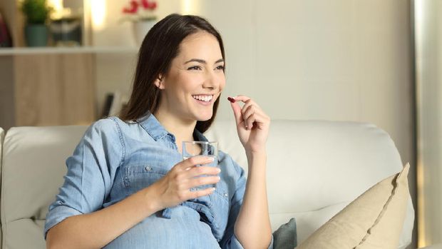 Portrait of a happy pregnant woman taking a pill sitting on a couch in the living room at home