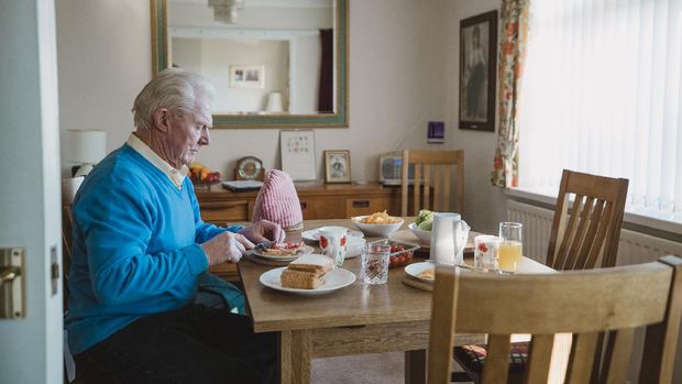 One senior man is sitting at the dining table in his home having lunch. He is making a ham salad sandwich.