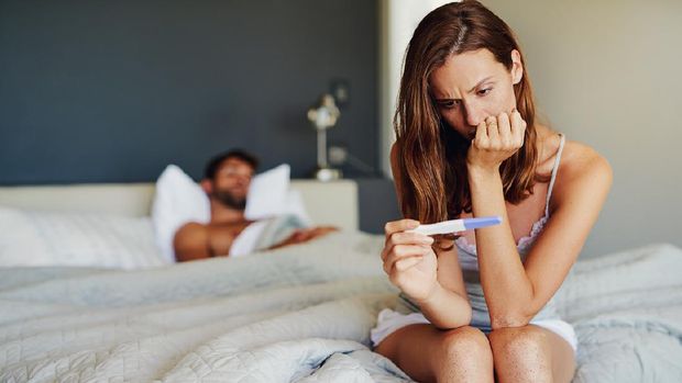 Shot of a woman looking worried while holding a pregnancy test with her partner lying in the background
