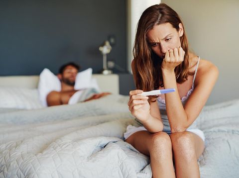 Shot of a woman looking worried while holding a pregnancy test with her partner lying in the background