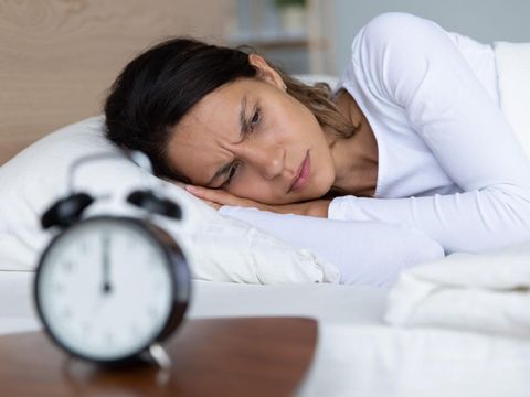 Unhappy restless woman wearing pajama suffering from insomnia, alarm clock on bedside table, sleepless tired girl with open eyes lying in bed, lack of sleep, early morning awakening concept