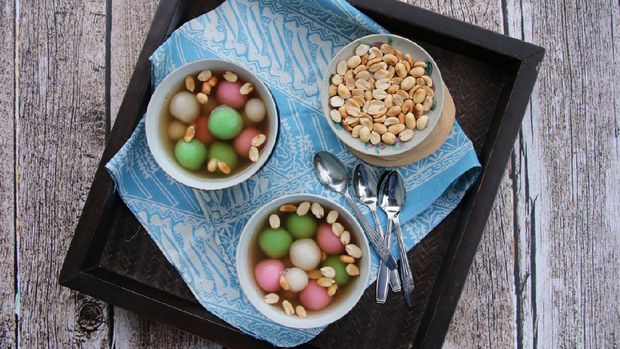 A Chinese Peranakan dessert of glutinous rice balls in warm ginger soup. Served in vintage ceramic bowls with extra peanuts, also in vintage saucer. This dessert dish is arranged on a wooden tray that has been lined with a batik napkin. The tray is then placed on a rustic wooden table.